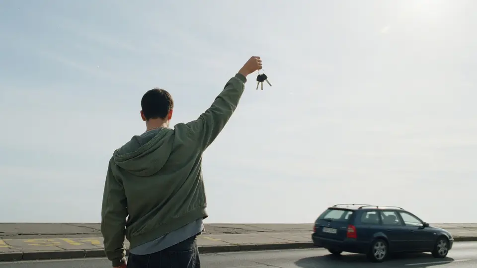 Jemand hält am Straßenrand einen Autoschlüssel hoch, im Hintergrund fährt ein Kombi vorbei.